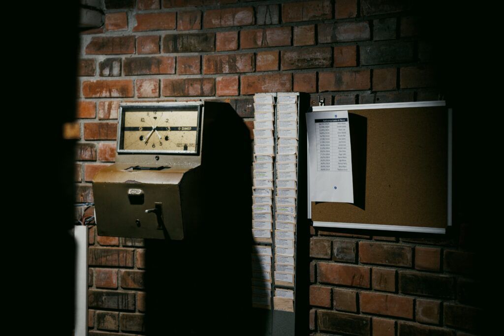 An old punch clock on a brick wall, next to a rack of time cards and a cork board with a sheet of paper pinned to it